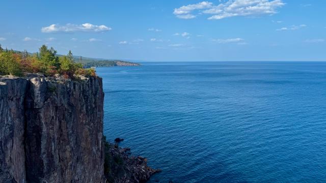 A vertical, rocky cliff overlooks a deep blue lake and verdant shores.
