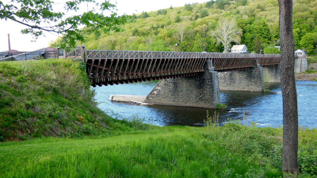 A wire suspension bridge crosses a river with a forested hill in the background.