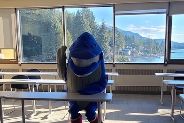 A whale mascot waves from a classroom seat overlooking a glacial bay